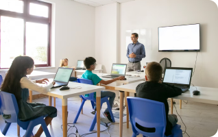 A teacher stands near a screen in a modern classroom, instructing children seated at computers.