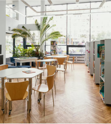 A library scene showcasing tables and chairs by a window, designed to foster economic efficiency in a study-friendly atmosphere.