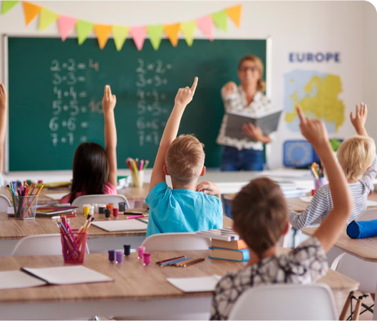 A group of children in a classroom, all raising their hands, indicating their eagerness to answer questions or engage in discussion.