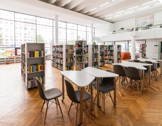 A library interior featuring tables and chairs arranged near large windows, emphasizing a well-maintained study environment.