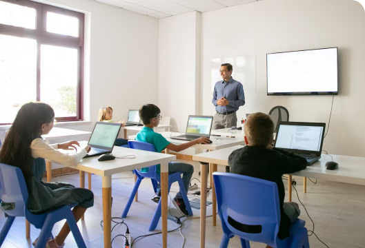 A man instructs children on computer usage, fostering their digital skills and encouraging learning in a collaborative environment.