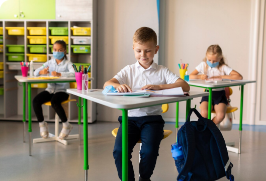 Children wearing masks sit at desks in a classroom, focused on their work and engaged in learning activities.