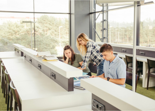 Three individuals collaborate at a library table, ensuring a safe and healthy workspace while engaged in their tasks.