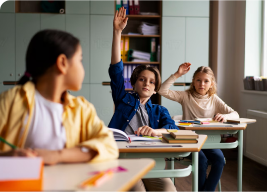 A group of children in an educational setting, seated at desks, eagerly raising their hands to participate.