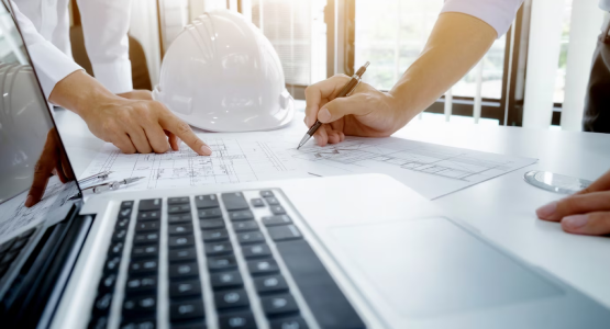 Construction workers in hard hats use a laptop together in a modular office setting on a job site.