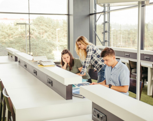 Three students collaborate at a library table, focusing on their studies in a well-ventilated indoor environment.