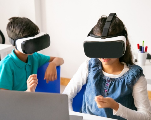 Two children engaged in virtual reality, wearing headsets while seated at a desk, exploring a digital world together.