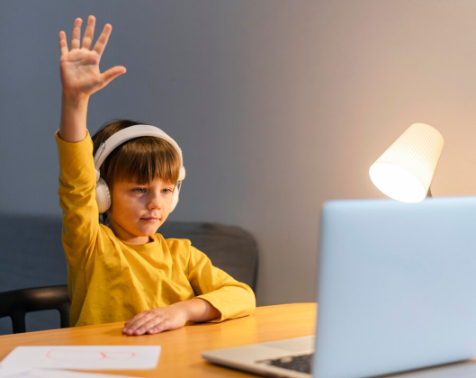 A young boy wearing headphones raises his hands at a desk, engaged in collaborative learning activities.