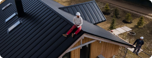 Two workers on a steep metal roof, one sitting near the ridge, the other standing on scaffolding.