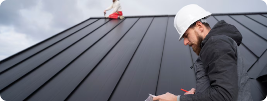 Person in hard hat examining paperwork with another individual working on a metal roof in the background.