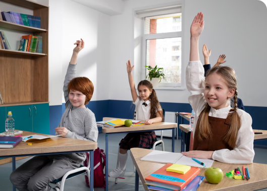 A group of children in a classroom, all raising their hands, demonstrating their eagerness to answer questions and engage.