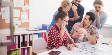 Several people gather at a table, engaged in discussion while reviewing a large blueprint.